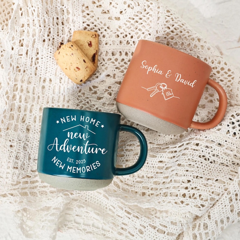 May include: Two ceramic mugs with a teal and orange exterior and a gray base. The teal mug has the text "NEW HOME, NEW ADVENTURE, NEW MEMORIES, EST. 2025". The orange mug has the names "Sophia & David" and a house and key graphic.