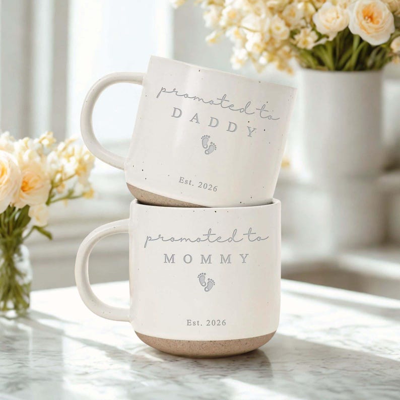 Op de afbeelding: Twee gestapelde witte keramische mokken met een gespikkelde afwerking. De bovenste mok leest "promoted to DADDY" met een voetafdruk en "Est. 2026". De onderste mok zegt "promoted to MOMMY" met een voetafdruk en "Est. 2026".