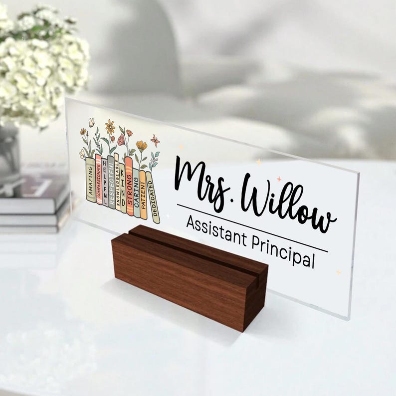 May include: A clear acrylic desk nameplate with the name "Mrs. Willow, Assistant Principal" in black script. The nameplate is supported by a dark wooden stand. Decorative elements include floral illustrations and a stack of books with positive adjectives.