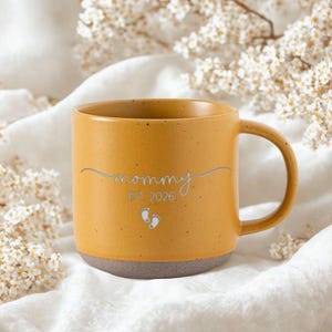 May include: Mustard yellow ceramic mug with a gray base. The mug features the word "mommy" in a silver script, the year "EST. 2026", and two small footprints. The mug has a curved handle.
