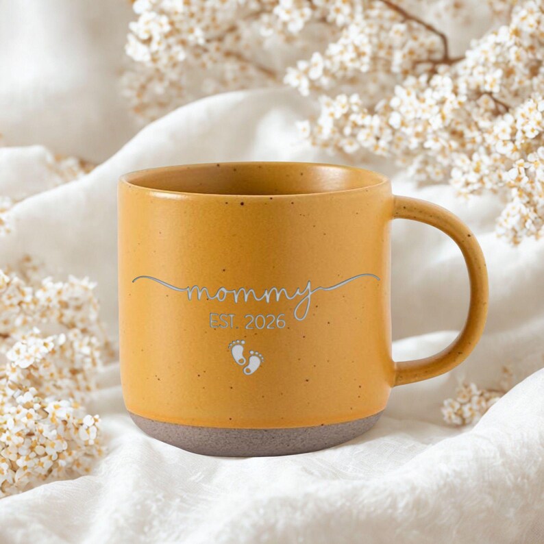 May include: Mustard yellow ceramic mug with a gray base. The mug features the word "mommy" in a silver script, the year "EST. 2026", and two small footprints. The mug has a curved handle.