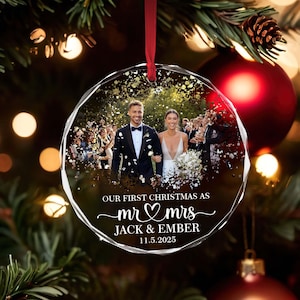 May include: A clear, faceted Christmas bauble featuring a wedding photo of a couple. The bauble has the text "OUR FIRST CHRISTMAS AS mr & mrs JACK & EMBER 11.5.2025" in white script. A red ribbon is attached.