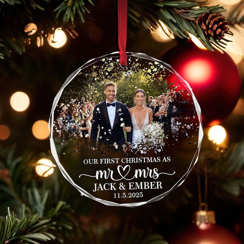 May include: A clear, faceted Christmas bauble featuring a wedding photo of a couple. The bauble has the text "OUR FIRST CHRISTMAS AS mr & mrs JACK & EMBER 11.5.2025" in white script. A red ribbon is attached.