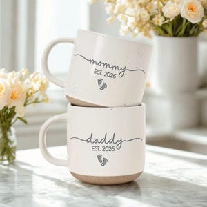 May include: Two stacked white ceramic mugs with a speckled beige base. The top mug says "mommy" with "EST. 2026" and baby footprints. The bottom mug says "daddy" with "EST. 2026" and baby footprints. The mugs have large handles.