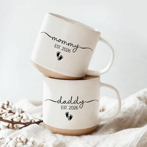 May include: Two stacked white ceramic mugs with a beige base. The top mug says "mommy EST. 2026" with a footprint graphic. The bottom mug says "daddy EST. 2026" with a footprint graphic. Both mugs have a curved handle.