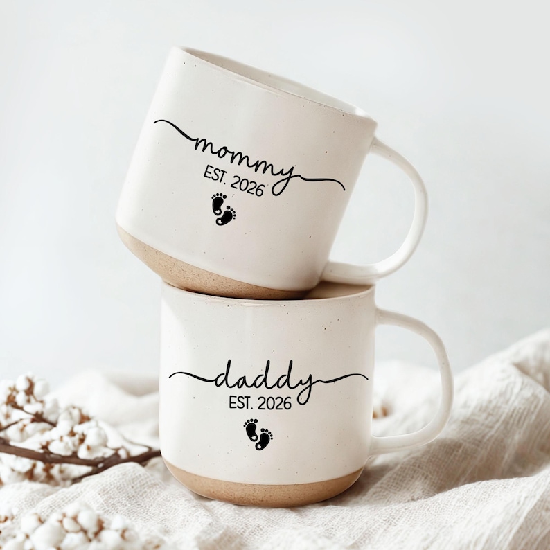 May include: Two stacked white ceramic mugs with a beige base. The top mug says "mommy EST. 2026" with a footprint graphic. The bottom mug says "daddy EST. 2026" with a footprint graphic. Both mugs have a curved handle.