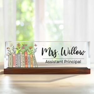 May include: A clear acrylic desk nameplate with a wooden base. The nameplate features the name "Mrs. Willow" and "Assistant Principal" in black script. Colorful books with positive adjectives and floral accents are on the left.