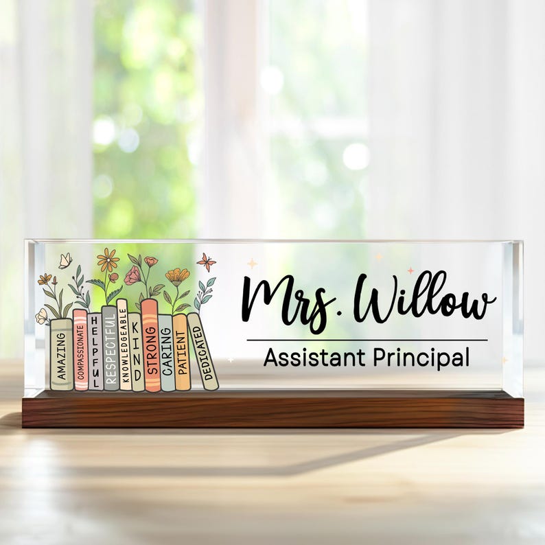 May include: A clear acrylic desk nameplate with a wooden base. The nameplate features the name "Mrs. Willow" and "Assistant Principal" in black script. Colorful books with positive adjectives and floral accents are on the left.