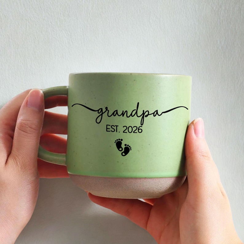 May include: A light green ceramic mug with the word "grandpa" in a cursive font, the year "EST. 2026", and two small black footprints. The bottom of the mug is a light beige color, and the handle is light green.