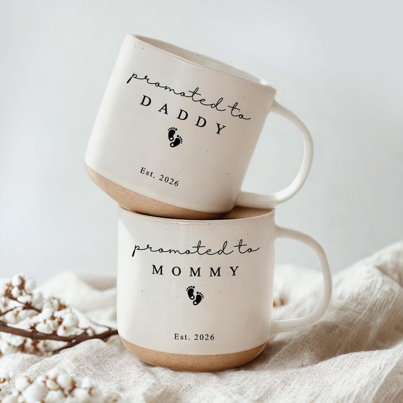 Op de afbeelding: Twee gestapelde witte keramische mokken met bruine bodems en handvatten. De bovenste mok leest "promoted to DADDY" met een voetafdruk en "Est. 2026". De onderste mok leest "promoted to MOMMY" met een voetafdruk en "Est. 2026".