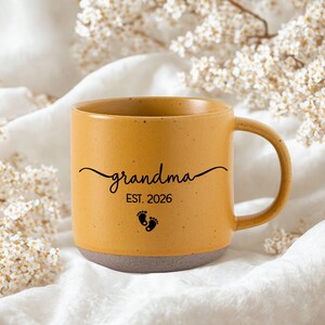 May include: Mustard yellow ceramic mug with a gray base. The mug features the word "grandma" in a cursive font, the text "EST. 2026", and two small footprints. The mug has a curved handle and is set against a soft, white background.