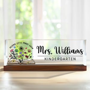 May include: A clear acrylic desk nameplate with a wood base. The nameplate features a colorful illustration of a book with flowers and butterflies. The text "Helping Little Minds Grow" is printed around the book. The nameplate also includes the text "Mrs. Williams Kindergarten".