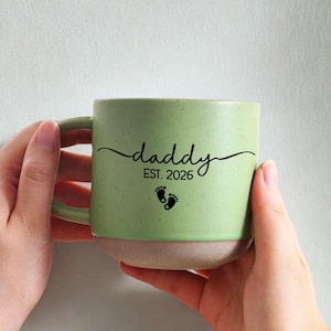 May include: A green ceramic mug with a light beige base. The mug features the word "daddy" in a script font, the text "EST. 2026", and two small black footprints. The mug has a rounded shape and a large handle.