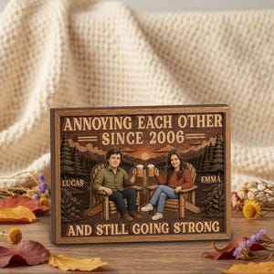 May include: A rectangular wooden sign featuring a printed illustration of two people sitting in chairs, holding beer mugs, with a mountain and forest background. The text reads: ANNOYING EACH OTHER SINCE 2006, LUCAS, EMMA, AND STILL GOING STRONG.