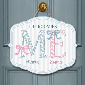 May include: A decorative wooden sign with the text "THE ROOMIES" above the letters "ME". The "M" is light blue with a floral pattern, and the "E" is pink with a floral pattern. The names "Maria" and "Emma" are below.