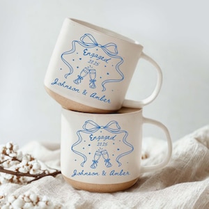 May include: Two stacked ceramic mugs with a cream-colored finish and a brown base. Each mug features a blue design with the word "Engaged" above the year "2025", a bow, two champagne glasses, and the names "Johnson & Amber".