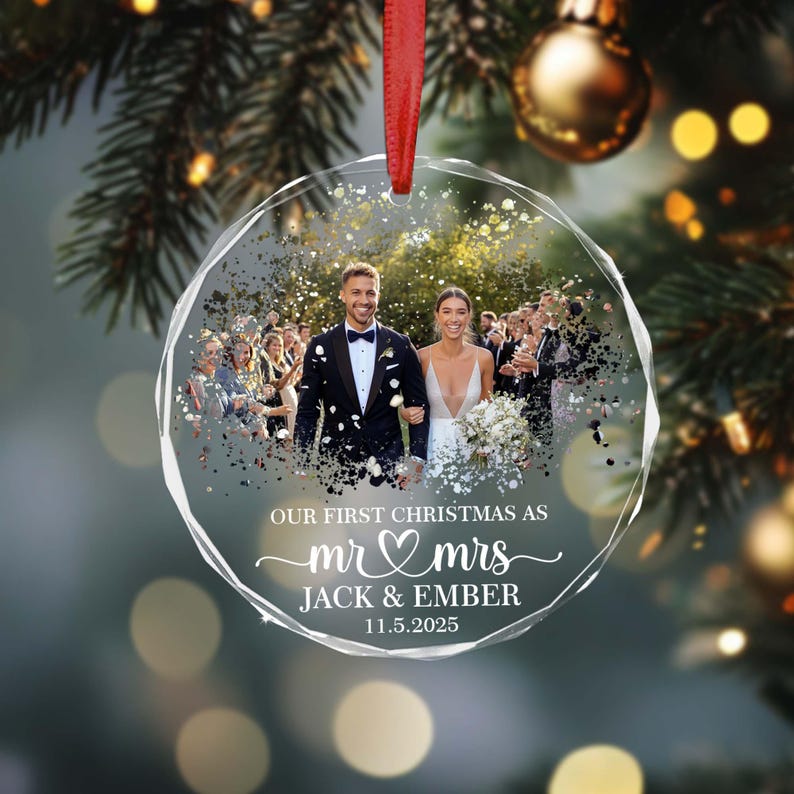 May include: Clear, faceted Christmas bauble with a wedding photo inside. The bauble has a red ribbon for hanging and features the text "OUR FIRST CHRISTMAS AS mr & mrs JACK & EMBER 11.5.2025". The background includes Christmas tree branches and gold baubles.