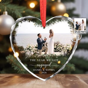 May include: A heart-shaped clear acrylic ornament featuring a printed photograph of a marriage proposal on a beach. The text reads THE YEAR WE GOT engaged DANIEL & SOPHIA 2026. A red ribbon is attached to the top for hanging on a tree.