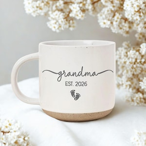 May include: Cream-colored ceramic mug with the word "grandma" in cursive, "EST. 2026", and baby footprints. The bottom of the mug is a light brown color. The mug has a curved handle and is surrounded by white flowers.