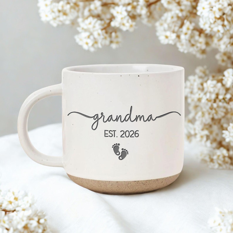 May include: Cream-colored ceramic mug with the word "grandma" in cursive, "EST. 2026", and baby footprints. The bottom of the mug is a light brown color. The mug has a curved handle and is surrounded by white flowers.