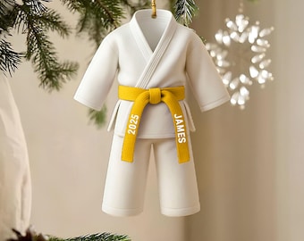 May include: A white karate uniform Christmas ornament with a yellow belt. The belt is inscribed with the name "JAMES" and the year "2025". The ornament is hanging from a gold string, set against a backdrop of a Christmas tree and a snowflake.