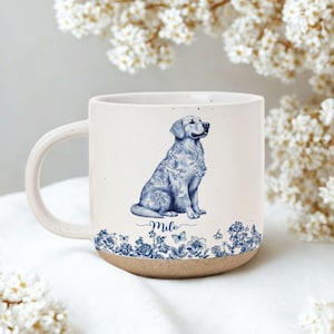 May include: A ceramic mug with a cream-colored body and a brown base. The mug features a blue and white illustration of a Golden Retriever dog with floral patterns. The dog's name, "Milo," is written in cursive below the dog.