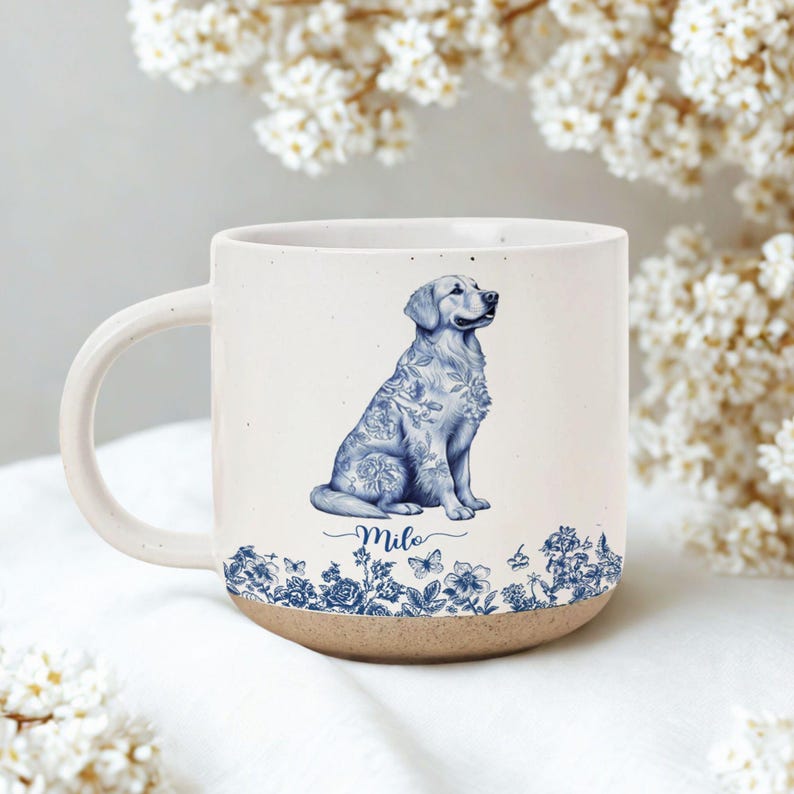 May include: A ceramic mug with a cream-colored body and a brown base. The mug features a blue and white illustration of a Golden Retriever dog with floral patterns. The dog's name, "Milo," is written in cursive below the dog.