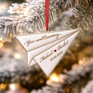 May include: A white paper airplane ornament with gold trim and a red ribbon hanger. The text reads 1 Year and Counting... Michael and Rachel with a small red heart between the names and the date 10.10.2025 engraved in gold lettering.