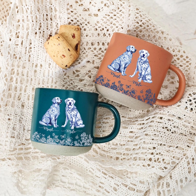 May include: Two ceramic mugs with dog illustrations. One mug is teal, the other is orange. Both mugs have a white floral design at the base and the names "Mush" and "Mile" below the dogs. The mugs are on a white lace surface.