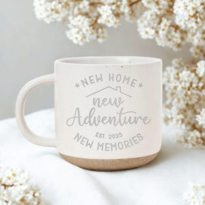 May include: Cream-colored ceramic mug with a light brown base and a curved handle. The mug features the text "NEW HOME, new Adventure, NEW MEMORIES EST. 2025" in a gray script. The mug is surrounded by white flowers.