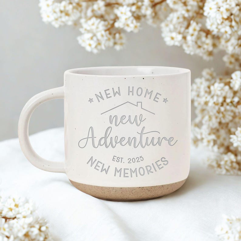 May include: Cream-colored ceramic mug with a light brown base and a curved handle. The mug features the text "NEW HOME, new Adventure, NEW MEMORIES EST. 2025" in a gray script. The mug is surrounded by white flowers.