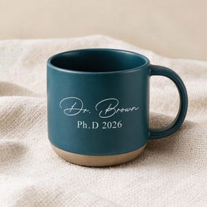 May include: A dark teal ceramic mug with a rounded base and a handle. The exterior features white text that reads Dr. Brown in a cursive script font, followed by the printed text Ph.D 2026. The bottom portion of the mug has a natural tan finish.