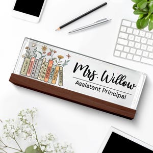 May include: A personalized desk nameplate with a dark wood base. The clear top displays the name "Mrs. Willow, Assistant Principal" in black script. Colorful books with positive adjectives and floral accents decorate the left side.