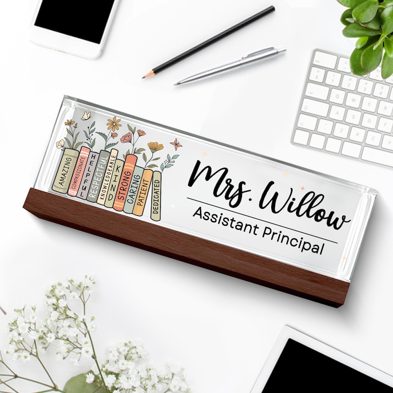 May include: A personalized desk nameplate with a dark wood base. The clear top displays the name "Mrs. Willow, Assistant Principal" in black script. Colorful books with positive adjectives and floral accents decorate the left side.