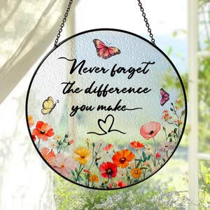 May include: Round glass suncatcher with a black metal frame and chain. The suncatcher features the text "Never forget the difference you make" with butterflies and colorful flowers. A decorative piece for a window.