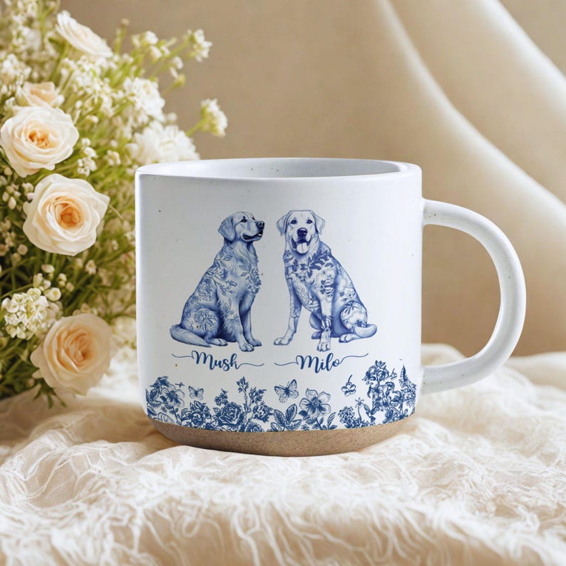 May include: White ceramic mug featuring two Labrador Retrievers with blue floral designs. The mug has the names "Mush" and "Milo" below the dogs. The bottom of the mug is a natural tan color.