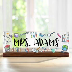 May include: A clear acrylic desk nameplate with a wooden base. The nameplate features a science theme with colourful illustrations of lab equipment, a microscope, a globe, and a book. The nameplate reads "MRS. ADAMS".