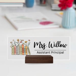 May include: A personalized desk nameplate with a dark wood base. The white sign features the name "Mrs. Willow" in elegant script, with "Assistant Principal" below. Colorful book spines with positive words and floral accents complete the design.