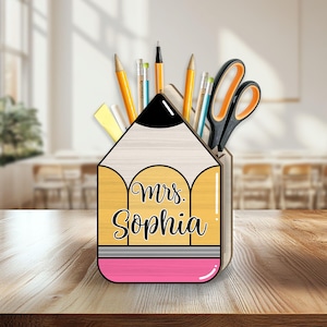 May include: A pencil-shaped desk organizer with the name "Mrs. Sophia" on the front. The organizer is filled with pencils, pens, scissors, and other stationery items. The pencil is yellow, black, white, and pink, and sits on a wooden surface.