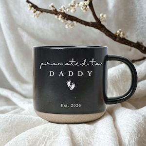 Op de afbeelding: Zwarte keramische mok met een beige basis. De mok heeft de woorden "promoted to DADDY" in witte letters, babyvoetjes en het jaar "Est. 2026". De mok staat op een witte, getextureerde ondergrond.