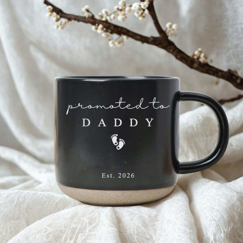 Op de afbeelding: Zwarte keramische mok met een beige basis. De mok heeft de woorden "promoted to DADDY" in witte letters, babyvoetjes en het jaar "Est. 2026". De mok staat op een witte, getextureerde ondergrond.
