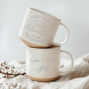 May include: Two stacked off-white ceramic mugs. The top mug has the text "NEW HOME, new Adventure, NEW MEMORIES, EST. 2025". The bottom mug has the text "Sophia & David" with a key and house graphic. Both mugs have a tan base.