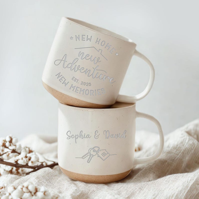 May include: Two stacked off-white ceramic mugs. The top mug has the text "NEW HOME, new Adventure, NEW MEMORIES, EST. 2025". The bottom mug has the text "Sophia & David" with a key and house graphic. Both mugs have a tan base.