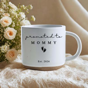 Op de afbeelding: Witte keramische mok met de tekst "promoted to MOMMY" in zwart schrift, babyvoetjes en het jaar "Est. 2026". De mok heeft een bruine basis en een wit handvat. De mok staat op een oppervlak met een patroon.
