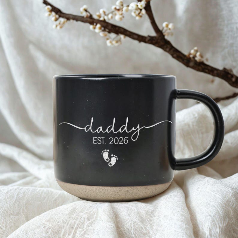 May include: Black ceramic mug with the word "daddy" in cursive, "EST. 2026", and baby footprints. The mug has a tan base and a black handle. The background is a white, textured fabric with a flowering branch.