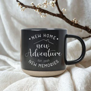 May include: Black ceramic mug with a beige base and a black handle. The mug features white text that reads "NEW HOME, NEW ADVENTURE, NEW MEMORIES, EST. 2025". A sprig of white flowers is in the background.