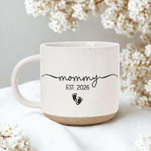 May include: Cream-colored ceramic mug with a brown base and a curved handle. The mug features the word "mommy" in a script font, the year "EST. 2026", and two small footprint graphics. The mug is set against a soft, floral background.