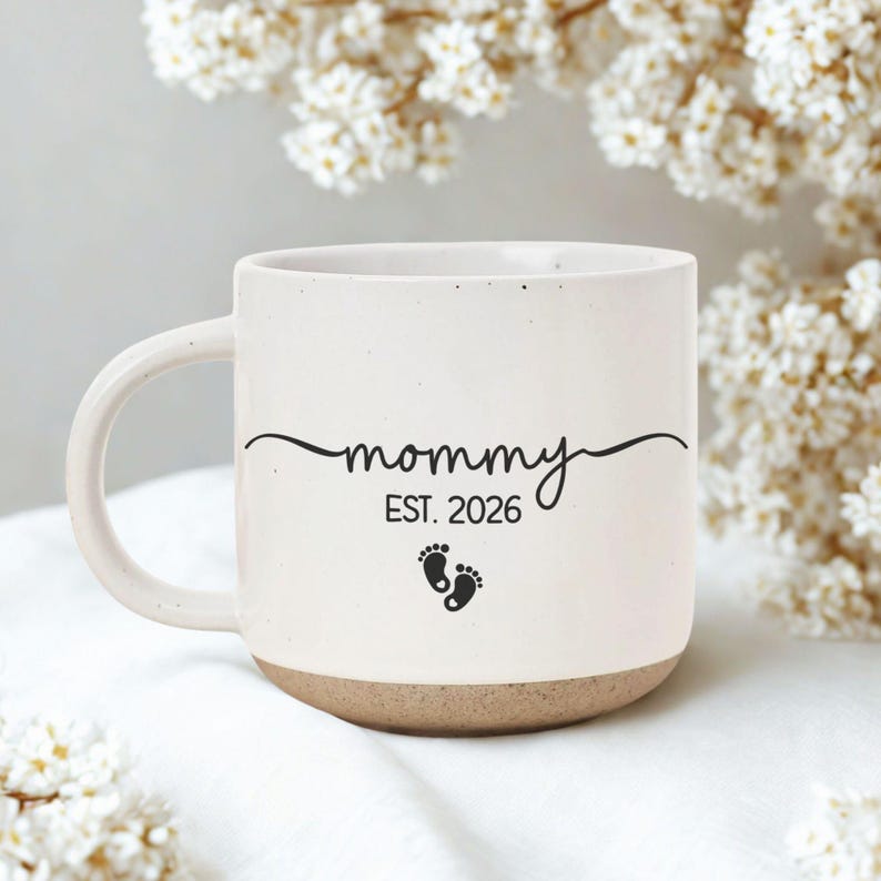 May include: Cream-colored ceramic mug with a brown base and a curved handle. The mug features the word "mommy" in a script font, the year "EST. 2026", and two small footprint graphics. The mug is set against a soft, floral background.
