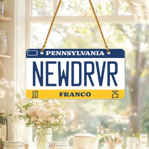 Puede incluir: Un letrero rectangular que se asemeja a una placa de matr&iacute;cula de Pensilvania, con el texto "PENNSYLVANIA" en la parte superior. La placa dice "NEWDRVR" en letras azules en negrita, con "10 FRANCO 25" en la parte inferior. El letrero cuelga de un cord&oacute;n dorado.