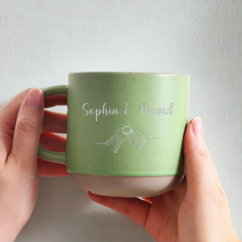 May include: A green ceramic mug with a light beige base. The mug features the names "Sophia & David" and a line drawing of keys and a house. The mug is held by two hands.